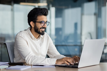 A smiling young Indian man in a suit sits in the office at a desk and works on a laptop, typing on...