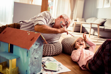 Grandfather and granddaughter painting a toy house together at home - obrazy, fototapety, plakaty