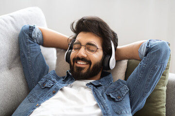 Content indian man wearing wireless headphones reclining peacefully on couch at home