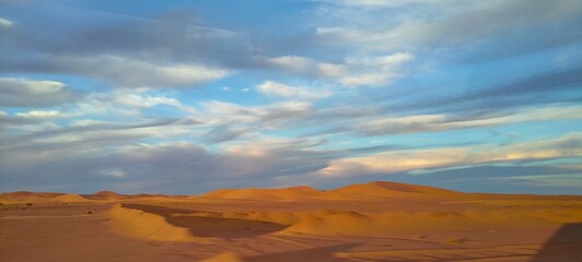 Naklejka premium A sweeping panorama captures the vast expanse of golden sand dunes stretching as far as the eye can see, forming an arid desert under a partly cloudy blue sky in Timimoun, Algeria.
