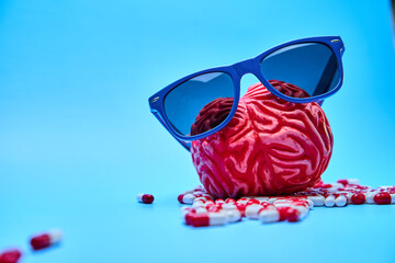 Red brain with sunglasses on top of a pile of red and white capsules on a blue surface.