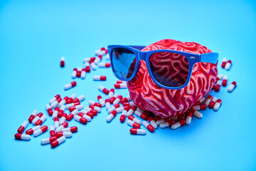 Red rubber brain with sunglasses on surrounded by white and red capsules on a blue surface. Concept of relaxation.