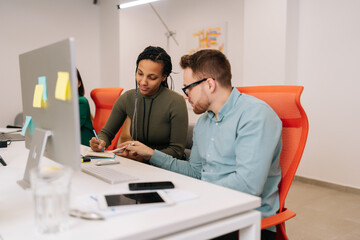 Two focused young diverse business people analysts partners talking, working together, discussing data management using desktop computer. Male manager consulting female client at office meeting.