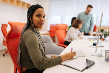 Portrait of beautiful African-American businesswoman writing down notes and using computer sitting...