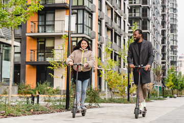 Young multiethnic couple riding electric scooters and talking on the street near modern buildings....