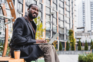 Smiling confident 30s african man using laptop with cup of coffee in city area. Young male...