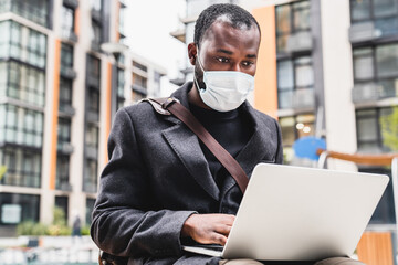 30s african man in mask against covid19 using his laptop in urban exterior. Businessman ceo manager...