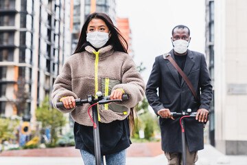 Multicultural couple on electric scooters ride together wearing masks to protect themselves from...
