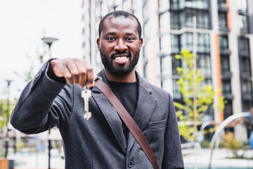 Happy smiling african 30s man holding keys of his new flat in front of modern block of flats...