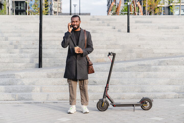 Busy african 30s man in elegant outfit talking on his phone close to electric scooter in city area. Businessman ceo manager wants to ride eco-transport to get to work office