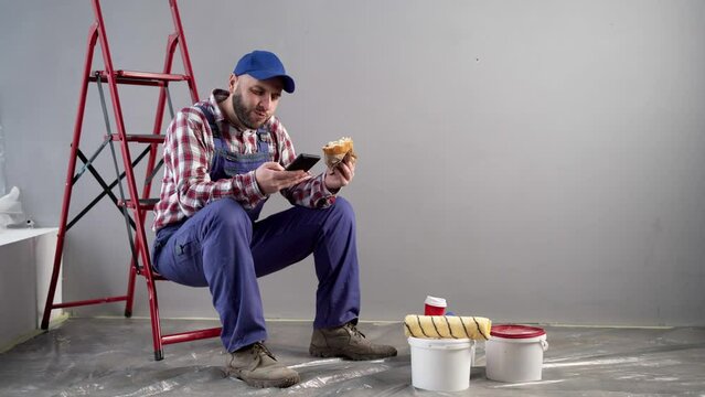 Professional painter having lunch with burger uses mobile phone for chatting with friends, sitting on ladder. Decoration and improvement home interior