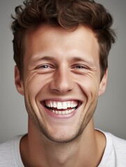 The image captures a portrait of a young man with short, curly hair and a very expressive, joyful smile, showcasing his white teeth. The background is a neutral, muted gray, and he is wearing a casual