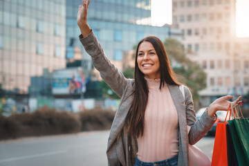 Beautiful young woman smiling widely while standing on the street and waving her hand in attempt to catch a taxi ride home.