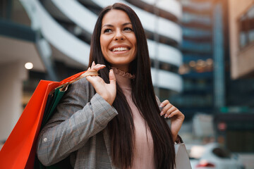 Portrait of pretty young woman with long straight hair standing in front of modern building and smiling while holding shopping bags.