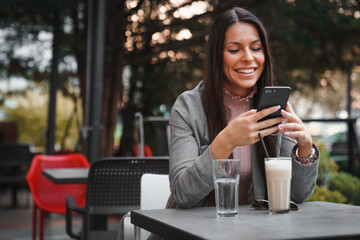 Attractive young woman sitting alone outdoors in a Cafe, enjoying her drink and texting while waiting a friend to join her.