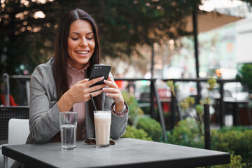 Happy young female entrepreneur sitting outdoors in a sidewalk Cafe, texting on her cellphone and smiling.