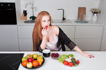 Young sexy woman eats ripe strawberries while sitting at the table in the kitchen. Healthy food. Diet.	