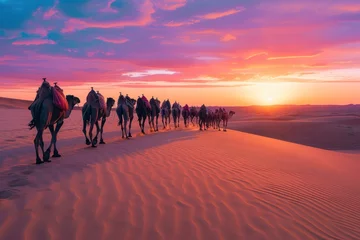  A caravan of camels walking in line on a desert dune under a vibrant sunset sky, leaving footprints in the sand. © Tuannasree