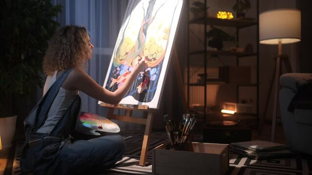 Beautiful Blonde Curly-Haired Female Painter. A Young Talented Artist wearing Headphones Draws a Picture and Enjoys Music while Sitting on the Floor in her Cozy Home Studio