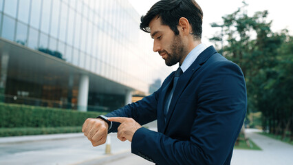 Skilled caucasian business man looking at time while standing at building. Attractive executive...