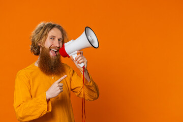 Caucasian young man talking with megaphone, proclaiming news, loudly announcing advertisement discounts sale, using loudspeaker to shout speech. Happy red hair guy isolated on studio orange background