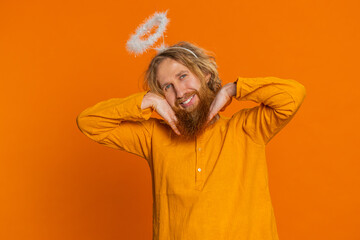 Portrait of smiling shy angelic Caucasian man with angel halo nimb over head flirting, looking at camera, positive emotions, celebrating holidays. Redhead young guy isolated on orange background