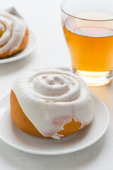 Two buns covered with white glaze on a saucer with a cup of tea on a light background.