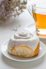 A bun covered with white glaze on a saucer, a cup of tea and a flower in a pot. Baking with tea in the morning light.