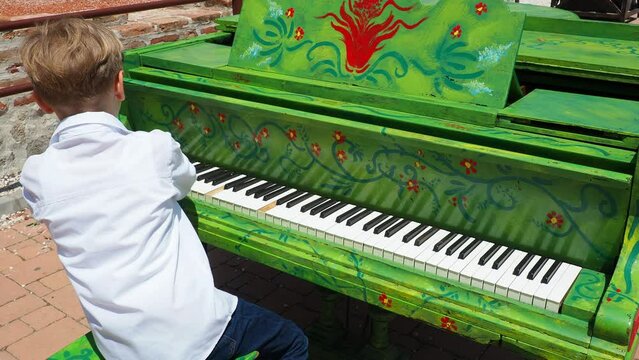 A blond boy in a white shirt plays the piano in the street. The child 9 years old Caucasian. Piano painted green. Musical performance. Street concert. Flash mob on the square. Musical excellence.