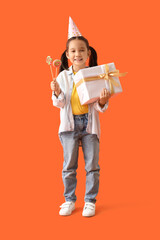 Cute little Asian girl in party hat with Birthday gift and lollipops on orange background