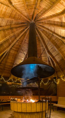 Big wooden grill house with beautiful wooden ceiling and giant grill in the middle with tables and glowing lights around