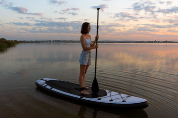 athletic woman on SUP paddle boarding through shining water at sunset. aesthetically wide shot....