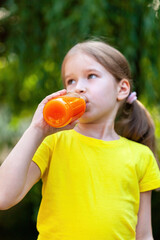 Young girl in a vibrant yellow t-shirt outdoors enjoying a refreshing drink from a clear bottle filled with orange carrot juice. Natural daylight, casual relaxed real lifestyle shot, vertical comp