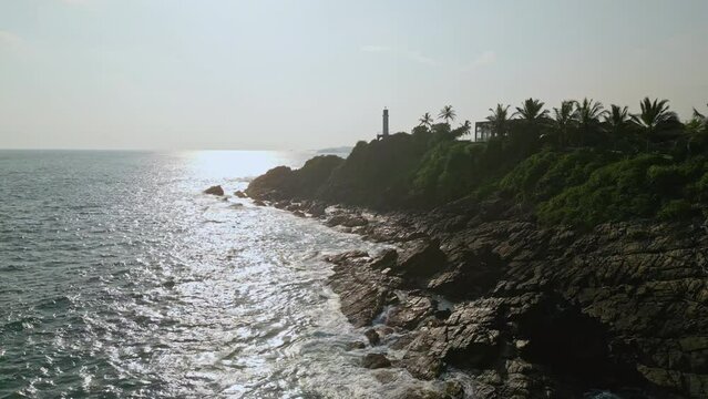 Aerial view of waves crashing on rocks near lighthouse, beacon for ships at sea, surrounded by lush foliage on hill, ocean navigation landmark, scenic coastal destination, sunny day shot.