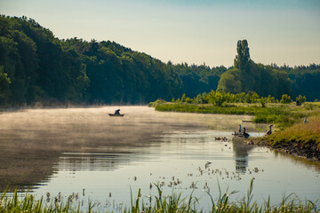 Summer scenery fisherman in a boat on a misty river at dawn ducks on the water