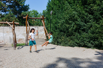 one girl playing and another one sitting on a pulley on a sunny day in the park