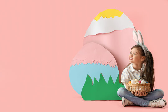 Cute little girl wearing bunny ears with basket of Easter eggs and decorations on pink background