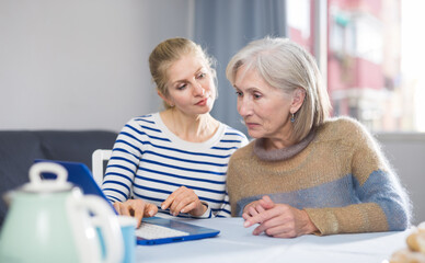 Mature woman and her adult daughter, sitting at a table, are viewing the necessary information on a...