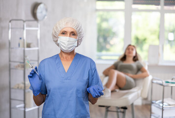 Portrait of trustworthy sneior doctor in scrub and mask holding syringe with patient lying in the back.