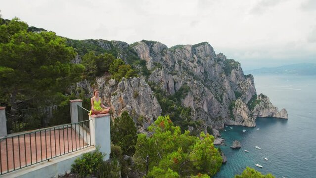 Person meditating on a balcony over Capri seaside cliffs at dusk. Woman peaceful posture against summer seascape in Italy.