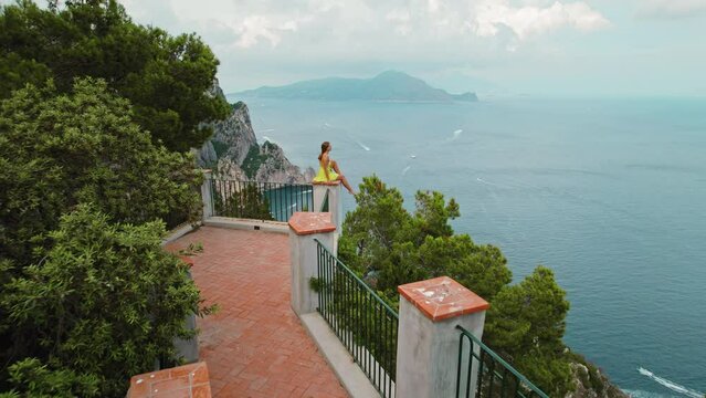 A woman in a yellow dress sits atop a scenic overlook. Girl gazes out at the serene seascape, where multiple boats dot the water's surface. Capri Italy.