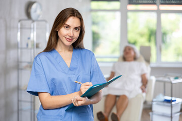 Young dentist in uniform holding documents folder in dental clinic with patient on couch behind