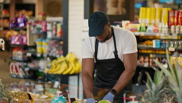 A man with Black skin in a white T-shirt and a Black apron using blue gloves manipulates goods on the counter in a large grocery store and his employee a man in a gray T-shirt, clarifies questions and