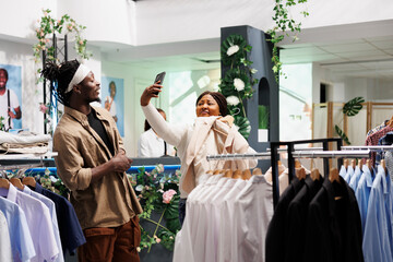 African american woman customer taking selfie on smartphone while choosing clothes in shopping mall. Fashion boutique clients trying formal wear and making photo on mobile phone