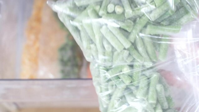 Female hand holds a plastic bag with frozen green beans on the background of freezer.