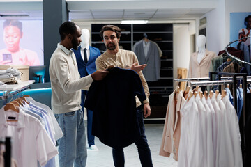 Fototapeta premium African american man holding shirt on hanger and asking shopping center worker about size options. Boutique worker pointing at new collection apparel rack, helping customer to choose outfit