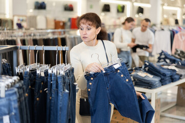 Watchful middle-aged female customer holding a hanger with dark jeans in large clothes store