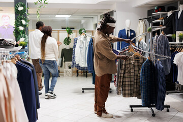 African american man searching outfit and choosing casual shirt on rack in shopping center....