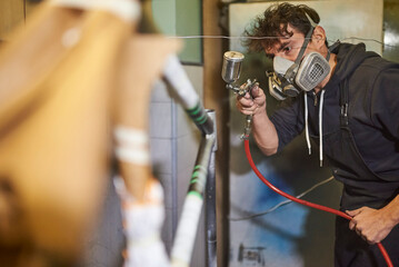 Latin young man wearing a protective respirator mask concentrated spray painting a bicycle frame in...