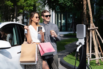 Young couple travel with EV electric car charging in green sustainable city outdoor garden in...